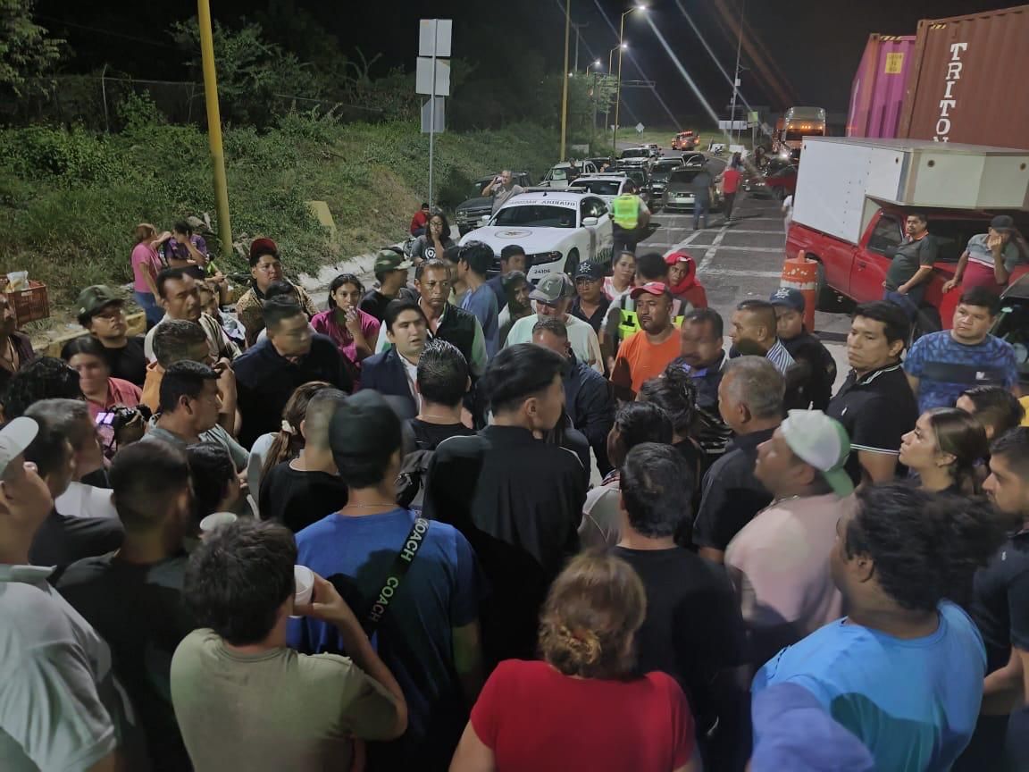 Diálogo nocturno con un grupo numeroso de personas sobre la autopista, con vehículos detenidos y contenedores de carga al fondo.