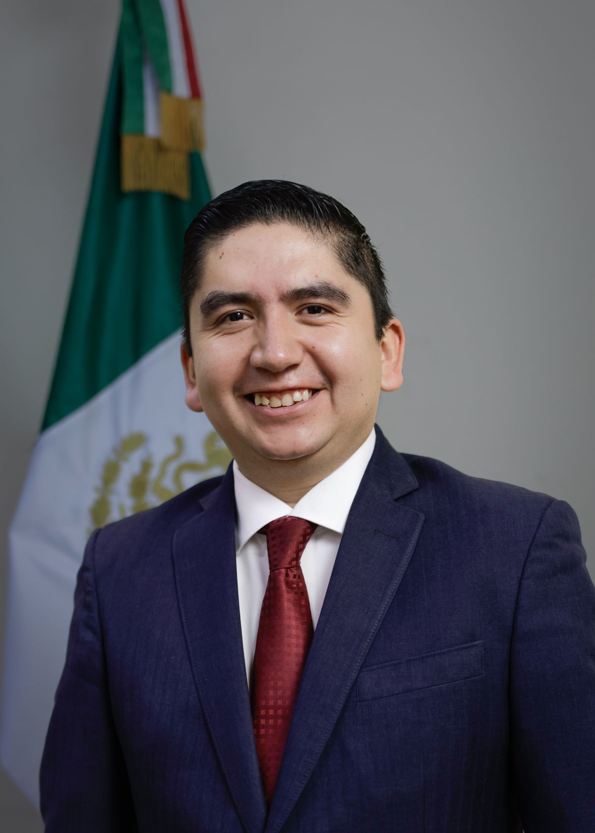 Retrato de Salvador J. Vazquez Villa, sonriente, con traje azul marino, camisa blanca y corbata roja.